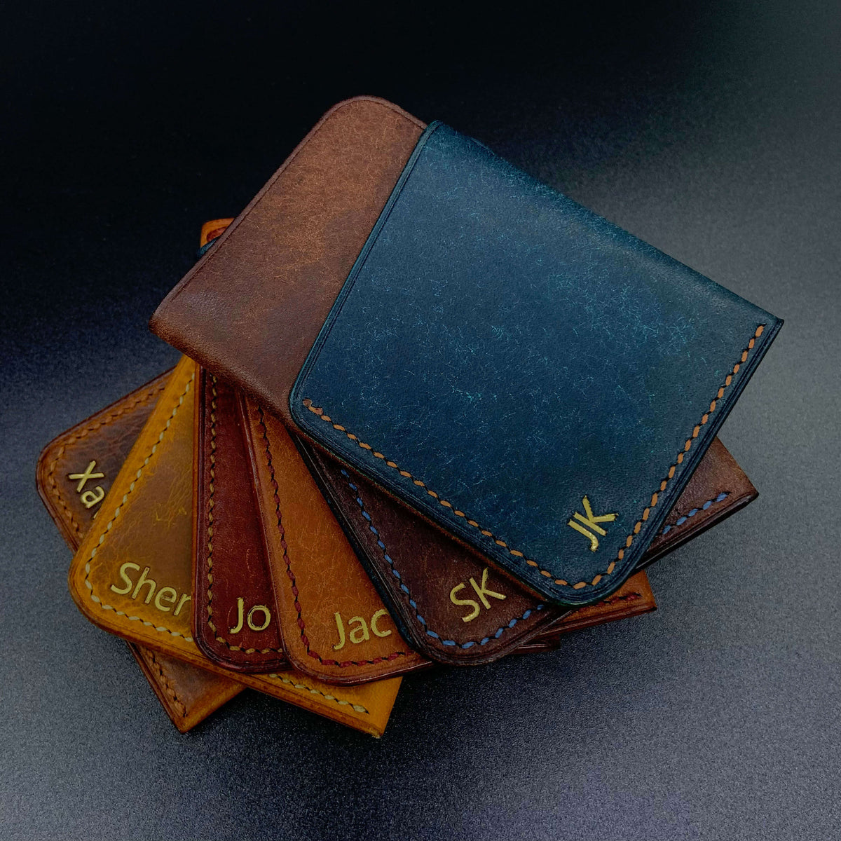 COZY Slim Sleeve Wallet personalised with gold foil names and initials in multiple colourways — tan, brown, navy and two-tone combinations fanned out on dark surface