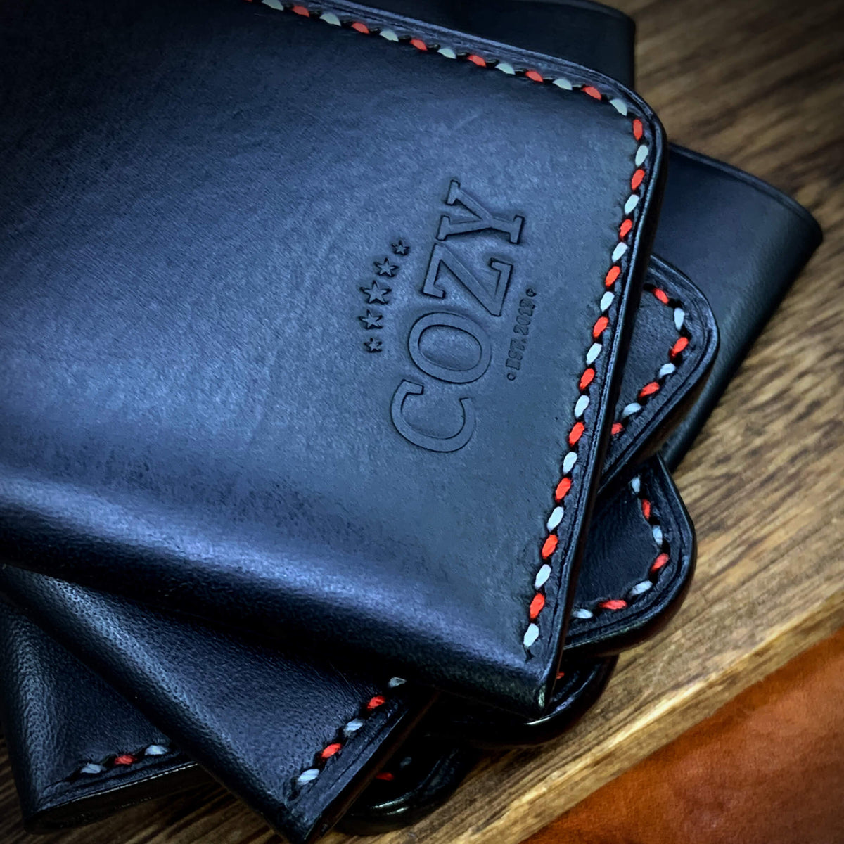 COZY Slim Sleeve Wallet SG Edition in pure black full grain leather stacked showing COZY stamp and red and white saddle stitch edge on wooden surface