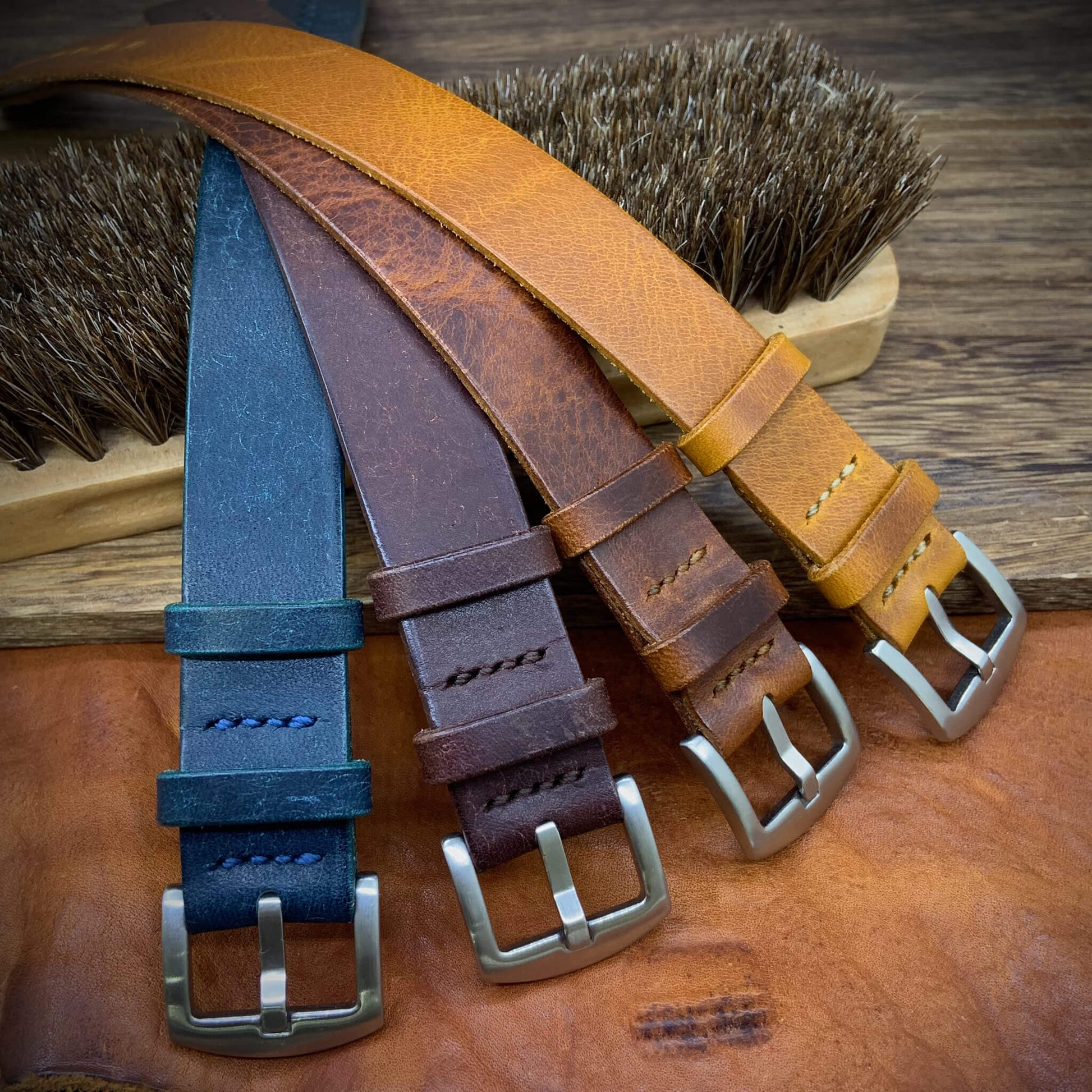 Four single-pass watch straps in different colors on a wooden surface with brushes in the background.
