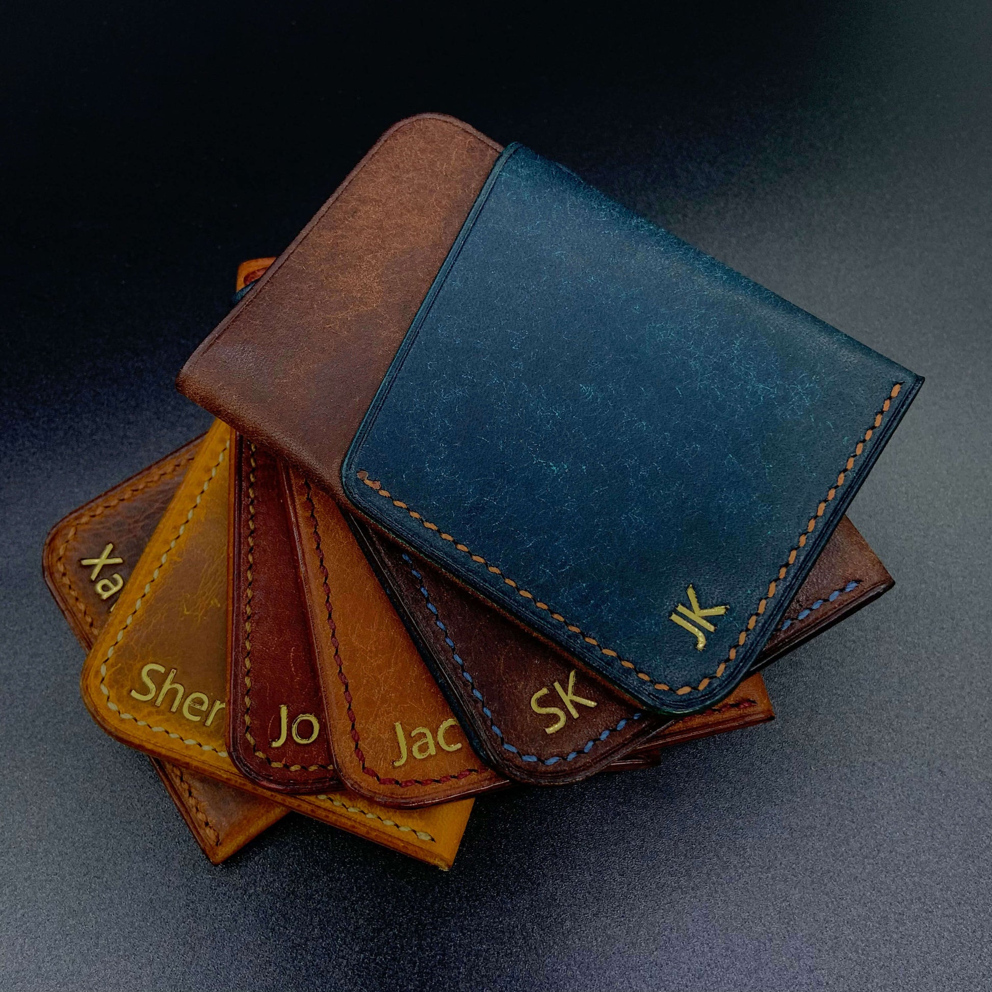 COZY Slim Sleeve Wallet personalised with gold foil names and initials in multiple colourways — tan, brown, navy and two-tone combinations fanned out on dark surface