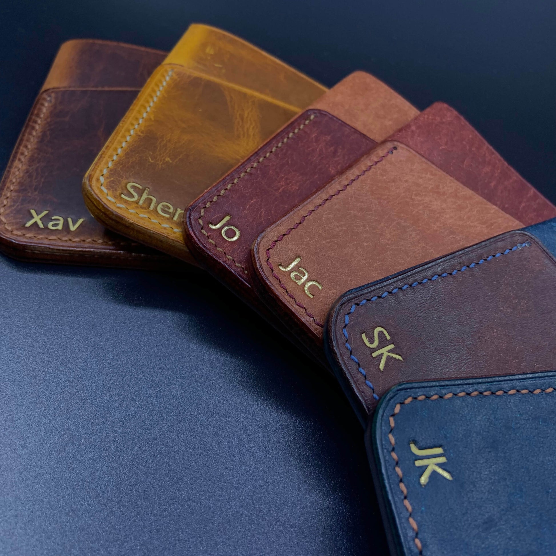 COZY Slim Sleeve Wallet personalised with gold foil initials and names fanned out on navy surface — showing Wax Cognac, Wax Napoli, Pueblo Tobacco and Pueblo Nazionale colourways with saddle stitch detail