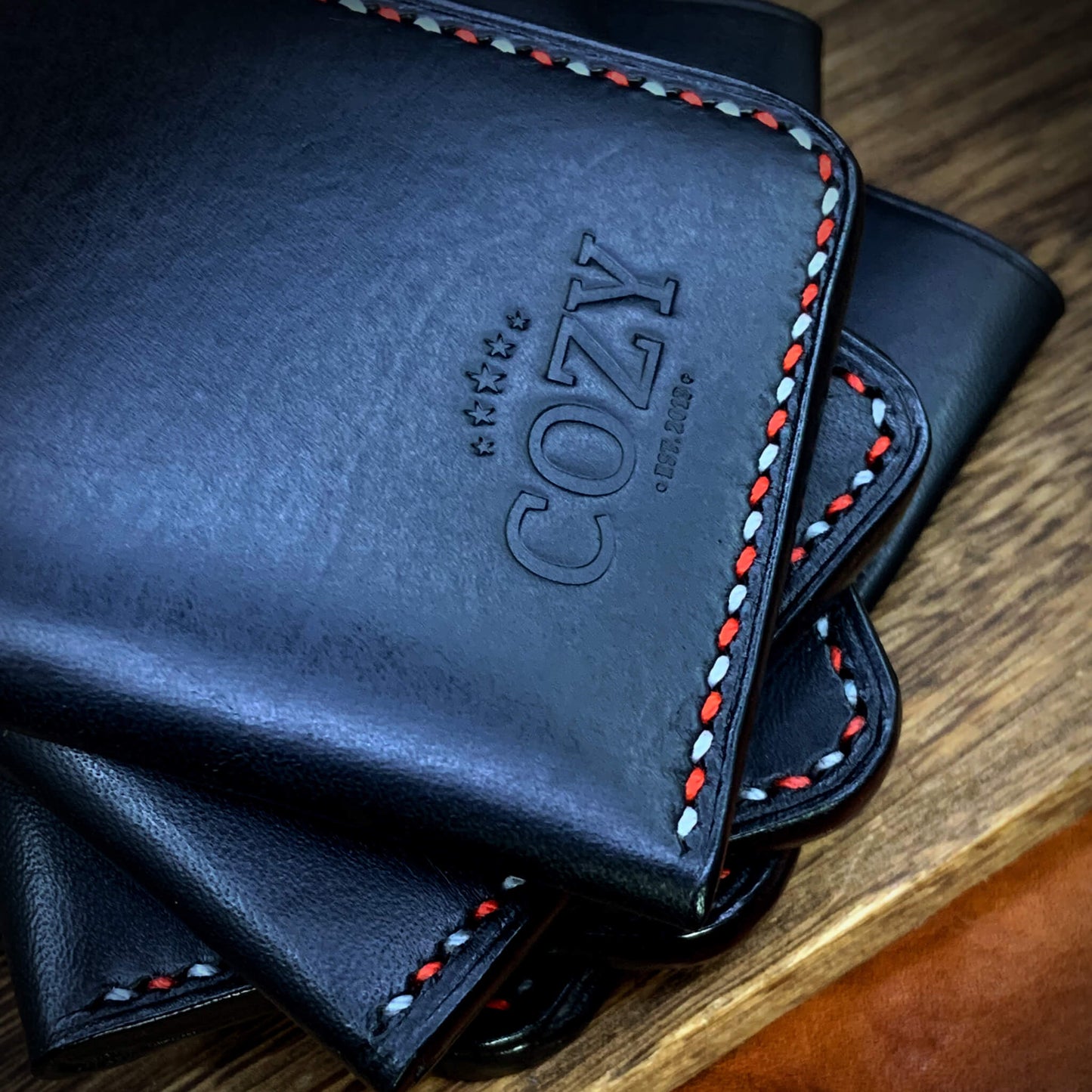 COZY Slim Sleeve Wallet SG Edition in pure black full grain leather stacked showing COZY stamp and red and white saddle stitch edge on wooden surface