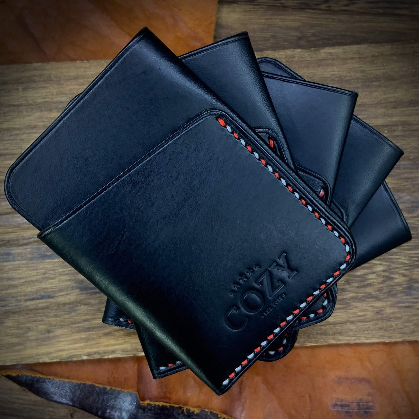 Multiple COZY Slim Sleeve Wallet SG Edition pieces fanned out on wooden surface — pure black full grain leather with alternating red and white saddle stitching