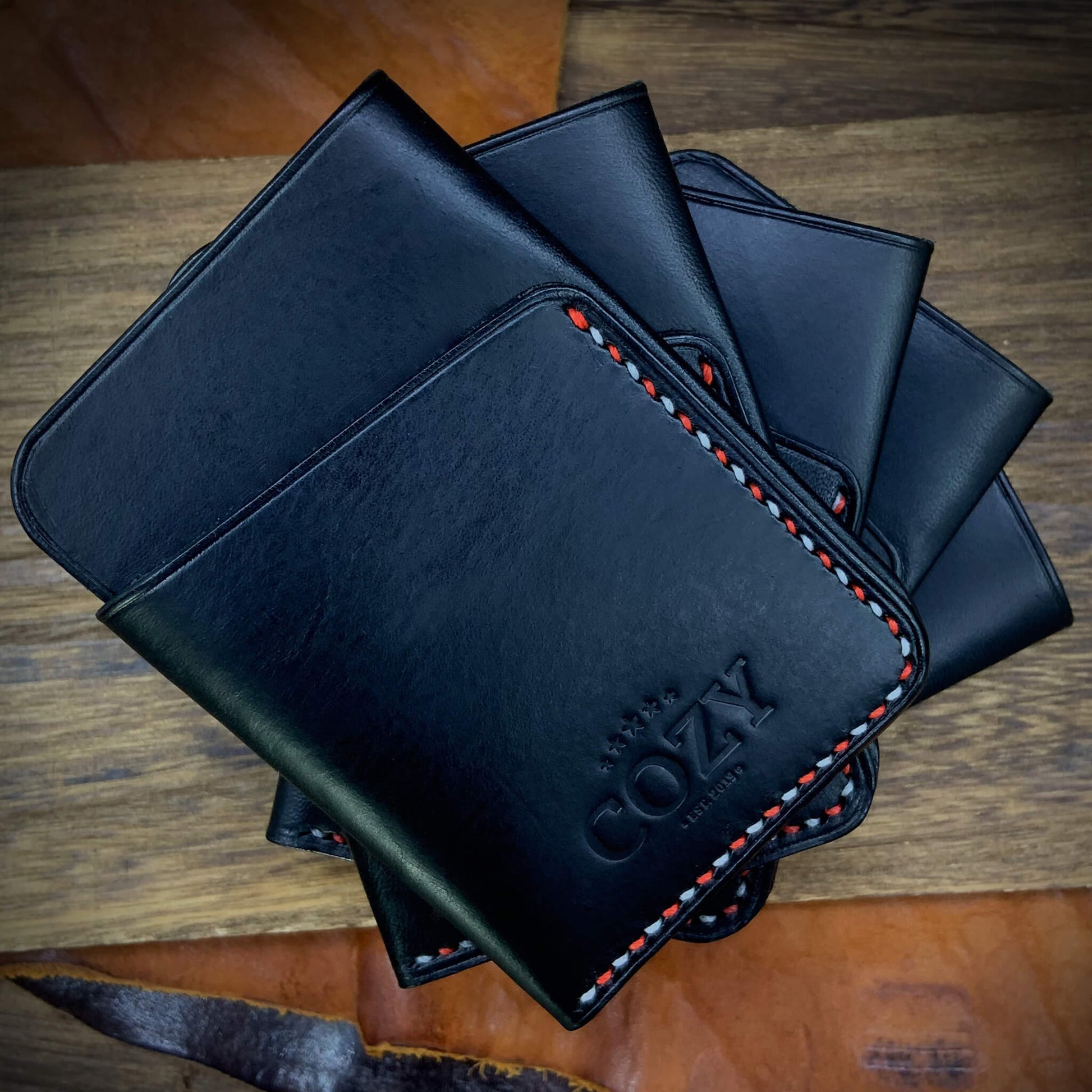 Multiple COZY Slim Sleeve Wallet SG Edition pieces fanned out on wooden surface — pure black full grain leather with alternating red and white saddle stitching