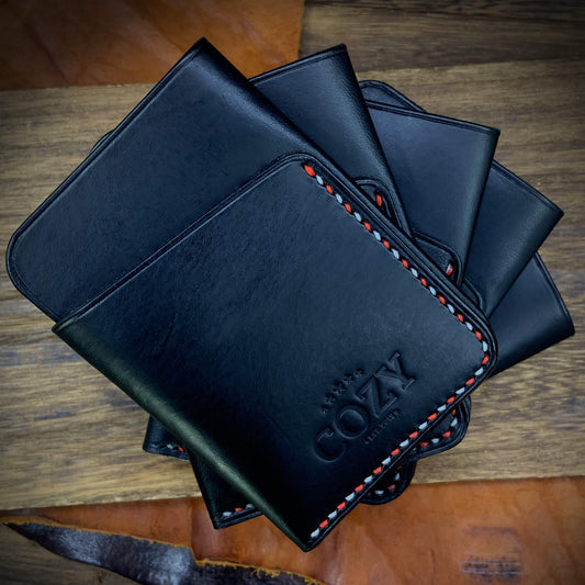 Multiple COZY Slim Sleeve Wallet SG Edition pieces fanned out on wooden surface — pure black full grain leather with alternating red and white saddle stitching