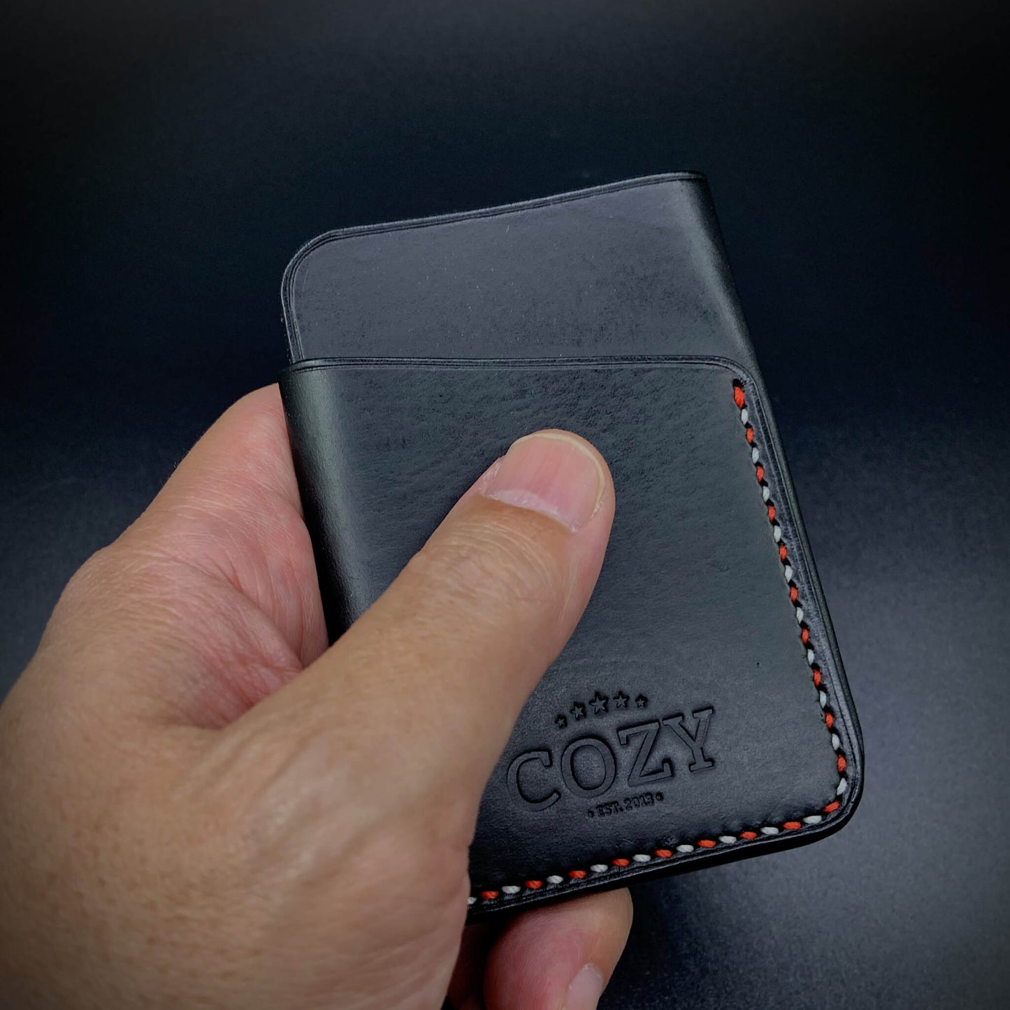 Hand holding COZY Slim Sleeve Wallet SG Edition in pure black full grain leather showing compact 7 x 10cm size and red and white saddle stitch detail