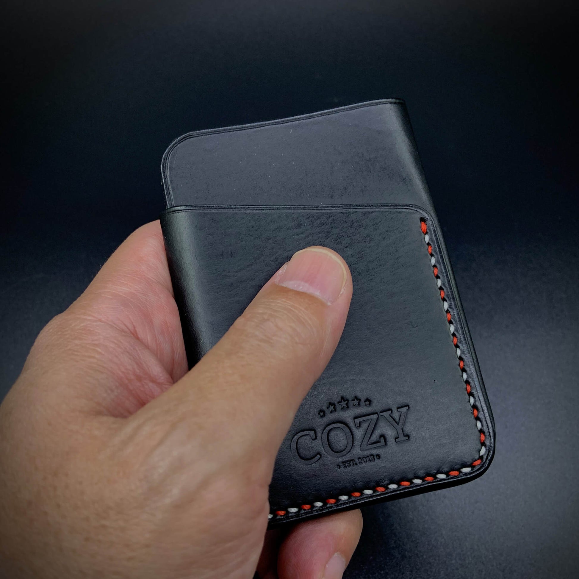Hand holding COZY Slim Sleeve Wallet SG Edition in pure black full grain leather showing compact 7 x 10cm size and red and white saddle stitch detail