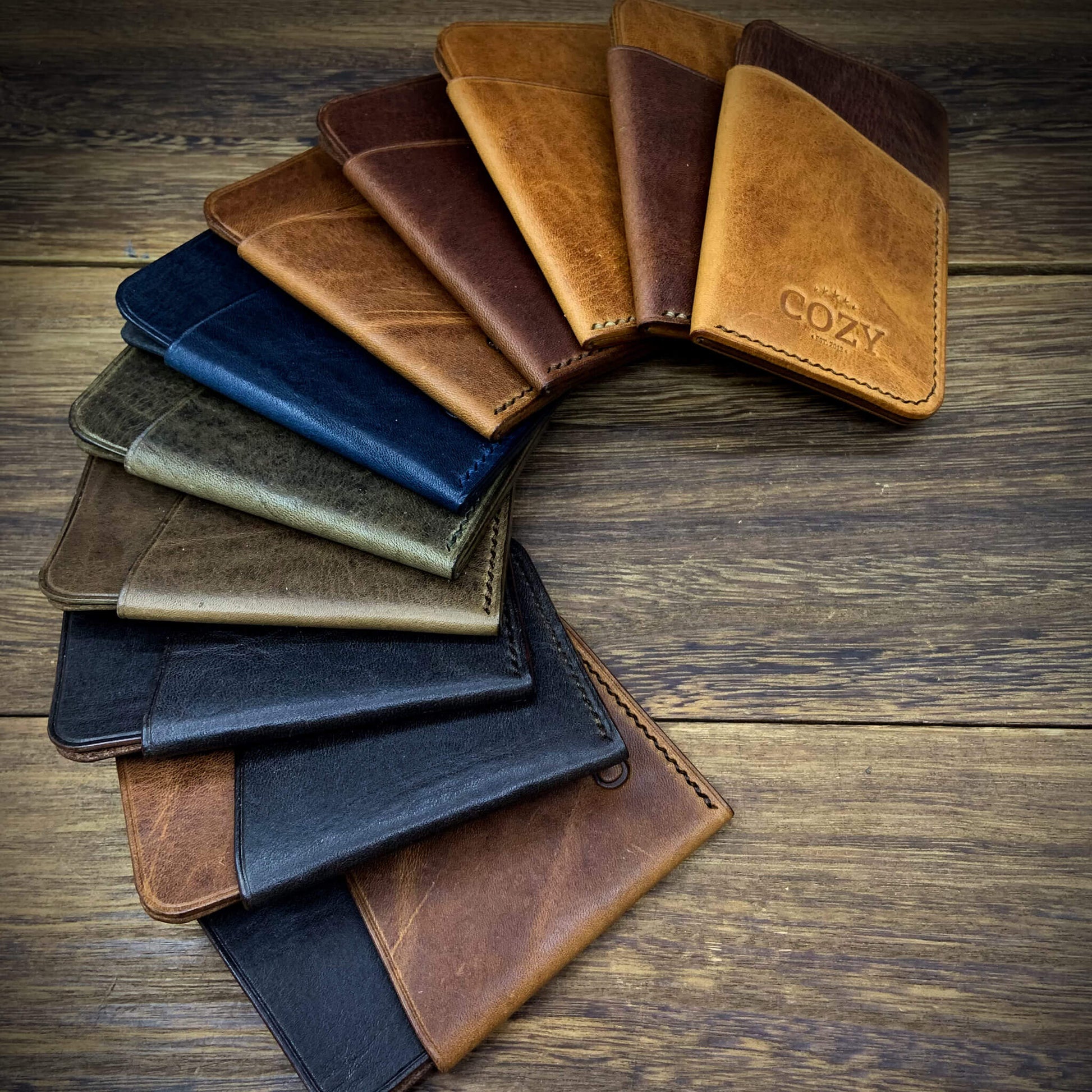 COZY Slim Sleeve Wallet in 11 colourways fanned out on wooden surface, showing full grain leather and hand-burnished edges